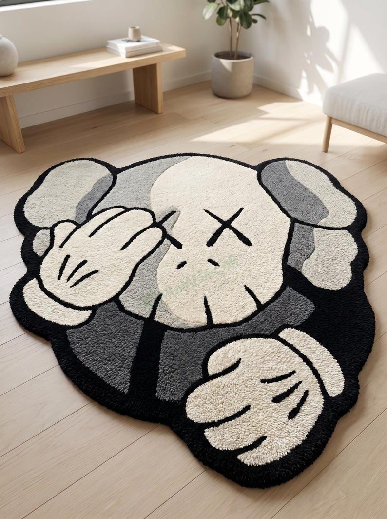 May include: A plush, character-shaped rug in shades of black, gray, and white. The rug features a cartoon-style design with a face and hands covering the eyes. The rug is placed on a light-colored wooden floor.
