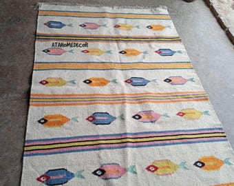 May include: A rectangular, off-white rug with a repeating pattern of colorful fish. The fish are in shades of yellow, orange, pink, and blue, with black eye details. Horizontal stripes in various colors separate the fish rows. The rug has fringed edges.
