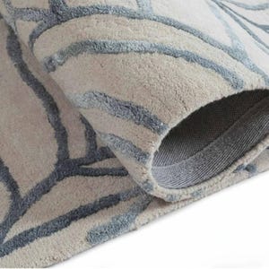 Puede incluir: Una alfombra enrollada con un fondo beige y un patrón geométrico azul. La textura de la alfombra es visible, y la parte inferior es de color gris sólido. El diseño presenta líneas curvas.