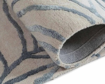 Può includere: Un tappeto morbido con una base color beige chiaro e un motivo in rilievo composto da linee fluide e interconnesse in un tenue blu ardesia. Il tappeto è ripiegato per mostrare il pelo denso e la struttura del supporto intrecciato.