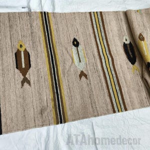 May include: Beige woven table runner with vertical stripes in black, brown, and yellow. The runner features embroidered fish motifs in brown, white, and yellow. The text "ATAhomedecor" is visible in the bottom right corner.