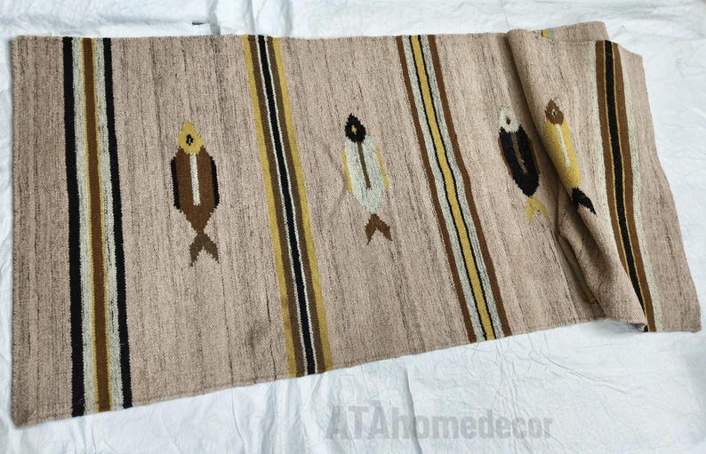 May include: Beige woven table runner with vertical stripes in black, brown, and yellow. The runner features embroidered fish motifs in brown, white, and yellow. The text "ATAhomedecor" is visible in the bottom right corner.