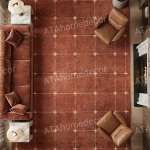 May include: A large rectangular area rug in a deep terracotta orange hue, featuring a grid pattern of thin white lines with small cross intersections. The rug is positioned in a living room setting with a brown sofa, two leather armchairs, and tables.