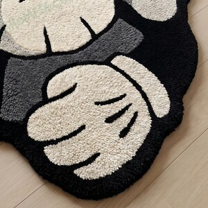 May include: A plush, cartoon-style rug in black, white, and gray. The rug features a hand design with a black outline and white, textured fingers. The background is black and gray, with a light-colored, textured surface.