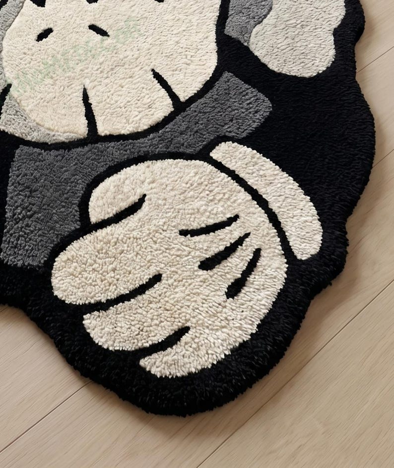May include: A plush, cartoon-style rug in black, white, and gray. The rug features a hand design with a black outline and white, textured fingers. The background is black and gray, with a light-colored, textured surface.
