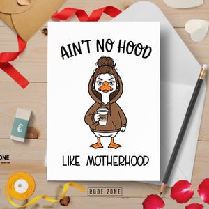 May include: A white greeting card with the text "AIN'T NO HOOD LIKE MOTHERHOOD" and a cartoon goose wearing a brown hoodie and holding a coffee cup. The card is on a wooden surface with a pencil, eraser, and red ribbon.