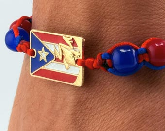 May include: A beaded bracelet featuring the Puerto Rican flag and a gold-colored frog charm. The bracelet has red, blue, and gold accents. The beads are approximately 0.5 inches in diameter.