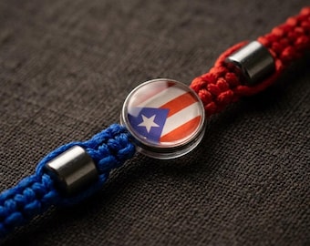 May include: A red and blue braided bracelet with a central charm featuring the Puerto Rican flag. The charm is round with a clear, domed surface. Silver-colored metal beads accent the bracelet.