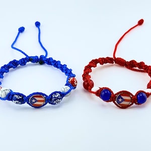 Puede incluir: Dos pulseras hechas a mano, una azul y otra roja, con nudos de macramé ajustables. Cada pulsera presenta cuentas y un dije central que muestra la bandera de Puerto Rico. La pulsera azul tiene cuentas blancas y rojas, mientras que la pulsera roja tiene cuentas azules y blancas.