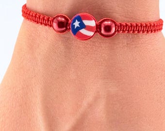 May include: Red braided bracelet featuring a central circular charm with the Puerto Rican flag design. The bracelet includes two red beads on either side of the charm. The bracelet is worn on a wrist.