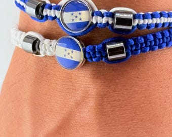 May include: Two bracelets with the flag of Honduras. One bracelet is white and the other is blue, both with a round emblem featuring the Honduran flag. The bracelets have a braided design and silver-colored metal accents.