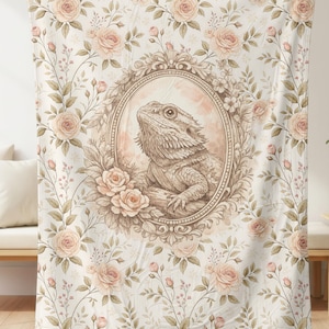 May include: A beige throw blanket featuring a detailed illustration of a bearded dragon within an ornate oval frame. The blanket is covered in a floral pattern of roses and leaves in shades of beige and pink.