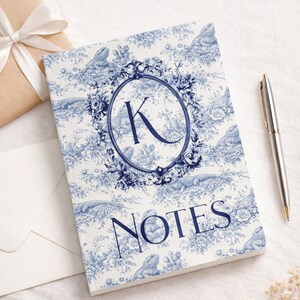 May include: A notebook with a blue and white toile pattern featuring lizards and floral elements. The cover includes the letter "K" inside an oval frame and the word "NOTES". A silver pen rests beside the notebook.