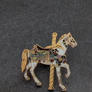 May include: A gold-toned metal enamel pin shaped like a carousel horse. The horse is white with a gold mane and tail, featuring a saddle decorated with a multi-colored floral pattern in shades of blue, red, and yellow, attached to a gold pole.