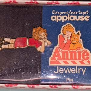 Może przedstawiać: Metalowa przypinka przedstawiająca postać z kręconymi rudymi włosami w czerwonej koszulce, zaprezentowana w pudełku z napisem Everyone loves to get applause Annie Jewelry Pin oraz ilustracją postaci i psa na niebieskim opakowaniu.