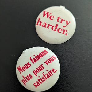 May include: Two circular white pin-back buttons with red text. One button features the text "We try harder." and the other button features the text "Nous faisons plus pour vous satisfaire." Both buttons have a small metal tab at the top edge.