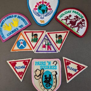 May include: A collection of embroidered fabric patches featuring various shapes and designs, including text such as International Tea '95, Holiday Parade, Cookies '95, Pairs 'n Squares, and Please, with colorful illustrations of people, globes, and symbols.