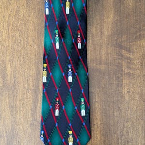 May include: A necktie featuring a dark navy blue and green diamond pattern overlaid with thin red and blue diagonal stripes. The tie is decorated with small, repeating illustrations of toy soldiers in various uniform colors including red, blue, green, and yellow.