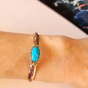 Puede incluir: Un brazalete de cobre con una piedra turquesa ovalada engastada en un bisel dorado. El brazalete tiene un diseño adornado.