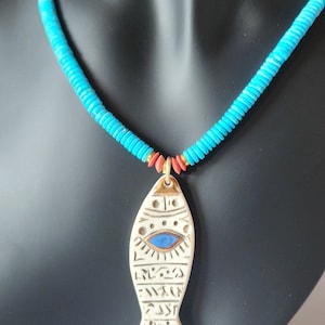 Puede incluir: Un collar de cuentas turquesa y rojo con un colgante de pescado de cerámica blanca. El pez tiene un ojo azul y detalles dorados.