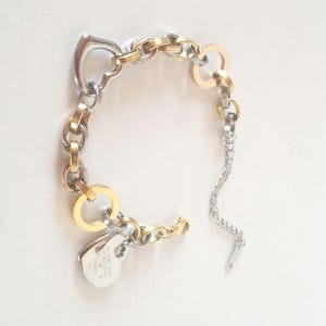 Pode incluir: Um bracelete de prata e ouro com um fecho em forma de coração e um amuleto com as palavras "Just be you, be kind, be true, be you" gravadas.