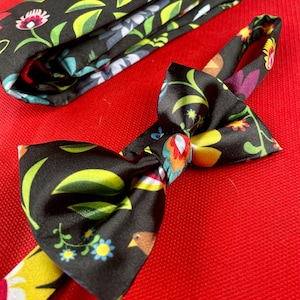 May include: A black bow tie with a colourful floral pattern. The bow tie is made of satin and has a yellow, red, green and blue floral design.