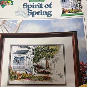 May include: A framed cross-stitch pattern of a white house with a porch and a garden. The title of the pattern is "Burton Dye's Spirit of Spring".