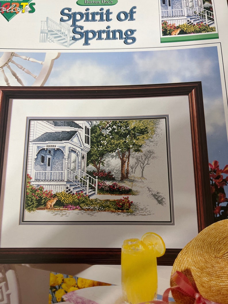 May include: A framed cross-stitch pattern of a white house with a porch and a garden. The title of the pattern is "Burton Dye's Spirit of Spring".