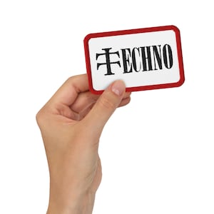 May include: A rectangular patch with a white background and a red border. The word "TECHNO" is printed in black, stylized lettering. The patch is held by a hand against a white background.