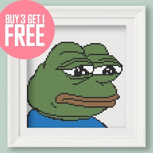 May include: A framed cross-stitch pattern of the popular internet meme "Feels Guy" or "Sad Frog". The frog is green with a blue shirt and has a sad expression on its face. The frame is white.