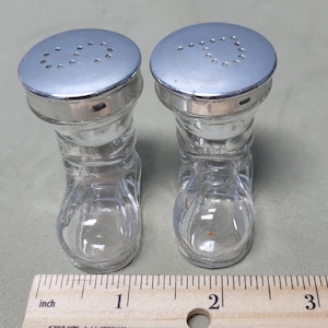 May include: A pair of clear glass salt and pepper shakers shaped like boots. Each shaker has a silver-colored metal lid with holes for dispensing. The shakers are displayed on a light green surface, with a ruler in the foreground.