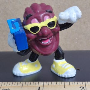 May include: A figurine of a smiling, anthropomorphic raisin wearing yellow sunglasses and sneakers. The raisin is holding a blue box and has a white glove raised in a fist. The figurine is approximately 2 inches tall.
