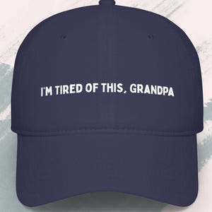 May include: A navy blue baseball cap with the text "I'M TIRED OF THIS, GRANDPA" in white. The hat has a curved brim and a structured crown. The brand name "CASUALTEES" is printed on the top left.