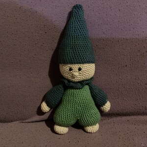 Pode incluir: Um gnomo de crochê verde com um chapéu pontudo. O gnomo tem um rosto bege com olhos pretos e um pequeno sorriso.