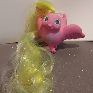 Op de afbeelding: Een roze speelgoedvogel met blauwe ogen en een lange, vloeiende geelgroene manen. De vogel heeft uitgestrekte vleugels en een klein, gedetailleerd ontwerp op de borst. Het is een grillig speelgoed.