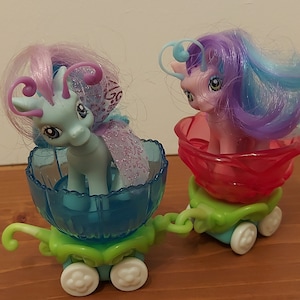 Puede incluir: Dos figuras de My Little Pony montando en carruajes con forma de flor. El pony de la izquierda es azul claro con pelo morado y rosa, y el carruaje es azul. El pony de la derecha es rosa con pelo morado y azul, y el carruaje es rojo.