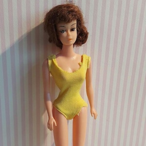 May include: A vintage doll with short, auburn hair and a yellow knit swimsuit. The doll has a slender figure and is posed against a pink and white striped background. The swimsuit has a scoop neckline and cap sleeves.