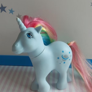 Op de afbeelding: Een lichtblauw eenhoorn pony speelgoed met een regenboog manen en staart. De pony heeft een zilveren hoorn en blauwe ogen. De pony heeft een blauwe en zilveren halve maan en sterren op zijn flank.