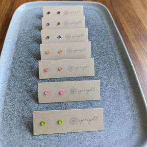 May include: Seven pairs of small, colorful stud earrings displayed on individual cards. Each card features the word "spright" and a small logo. The earrings are in the colors orange, pink, green, blue, and purple.