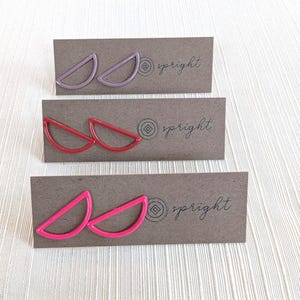 May include: Three pairs of earrings in shades of pink, red, and purple, each in a half-circle design. The earrings are displayed on brown cards with the word "spright" printed on them. The background is a light, textured surface.