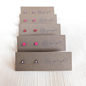 May include: A collection of stud earrings in various colors, including pink, red, and purple, displayed on individual brown cardboard cards. Each card is printed with the word "spright" and a circular logo.