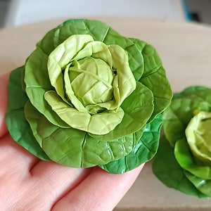 May include: Two green cabbage heads made of polymer clay. The cabbage heads are arranged in a bouquet-like shape.