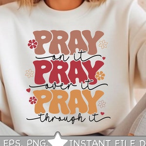 Puede incluir: Una sudadera blanca con el texto "Pray on it, Pray over it, Pray through it" en una fuente retro colorida. El texto está rodeado de pequeñas flores y corazones.