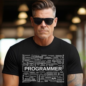 May include: A black t-shirt featuring a white word cloud design that reads "PROGRAMMER". The word cloud includes terms related to software development, such as "frontend", "backend", "codebase", "debugging", "algorithms", "version control", "API", and "mobile app development".