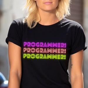 Pode incluir: Uma mulher vestindo uma camiseta preta com o texto "PROGRAMMER!" repetido três vezes em um degradê de rosa, laranja e amarelo. Ela está sentada em uma superfície de concreto com uma motocicleta ao fundo.