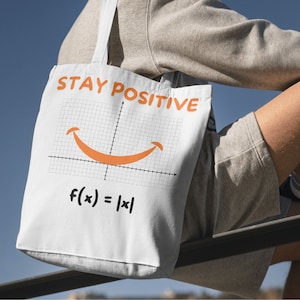 May include: White tote bag with the words "STAY POSITIVE" in orange, and a smiling graph design. The equation f(x) = |x| is printed below the graph. The bag has white straps and is made of a durable fabric.