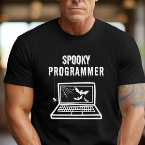 Può includere: T-shirt nera con la scritta "SPOOKY PROGRAMMER" sopra una grafica di un laptop con una ragnatela, pipistrelli e ragni. La persona nella foto indossa occhiali da sole.