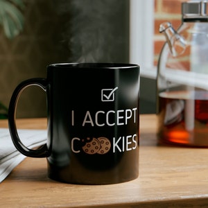 Könnte beinhalten: Schwarze Keramik-Kaffeetasse mit weißem Text, der "Ich akzeptiere Cookies" lautet und einem Häkchen. Zwei braune Schokoladenkekse sind neben dem Wort "Cookies" abgebildet.
