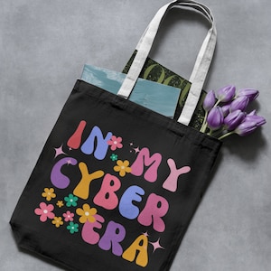 May include: Black tote bag with white handles featuring the phrase "IN MY CYBER ERA" in colorful, retro-style lettering. The design includes flowers and stars. The bag is filled with books and purple tulips.