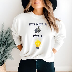 May include: White sweatshirt with a graphic of a bug and a lightbulb. The text on the sweatshirt reads "It's not a" above the bug and "It's a" below the lightbulb.